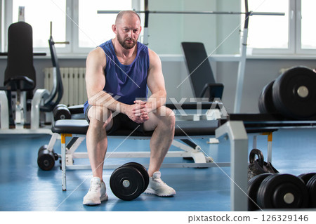 A man is sitting on a bench in a gym. He looks tired and depressed, nothing makes him happy. A man is sitting on a bench in a gym. He looks tired and depressed, nothing makes him happy. 126329146