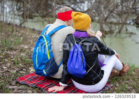 Active elderly Caucasian couple hiking with backpacks and hiking poles enjoying their adventure. They sit with their backs to the camera. Active elderly Caucasian couple hiking with backpacks and hiking poles enjoying their adventure. They sit with their backs to the camera. 126329518