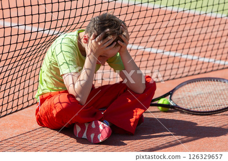 The boy lost the tennis competition. The child sits at the net with a racket. Psychology of sports in childhood. The boy lost the tennis competition. The child sits at the net with a racket. Psychology of sports in childhood. 126329657