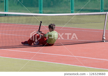 The child is sitting at the tennis net with a racket. He lost the competition or is tired of training. The child is sitting at the tennis net with a racket. He lost the competition or is tired of training. 126329660
