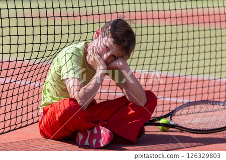 The boy lost the tennis competition. The child sits at the net with a racket. Psychology of sports in childhood. The boy lost the tennis competition. The child sits at the net with a racket. Psychology of sports in childhood. 126329803
