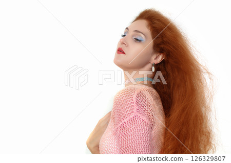 A woman with red hair and blue eyes stands in front of a white background. She is wearing a pink sweater and blue earrings A woman with red hair and blue eyes stands in front of a white background. She is wearing a pink sweater and blue earrings 126329807