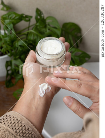 Woman touching cream with finger and holding a cosmetic jar in bathroom against green plant Woman touching cream with finger and holding a cosmetic jar in bathroom against green plant 126330287
