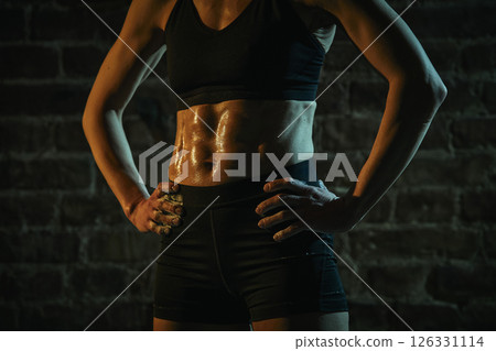 Cropped photo. Woman stands in gym with hands on hips and tight abs glistening with sweat, radiating strength, resilience and post-workout pride under dramatic light. Cropped photo. Woman stands in gym with hands on hips and tight abs glistening with sweat, radiating strength, resilience and post-workout pride under dramatic light. 126331114