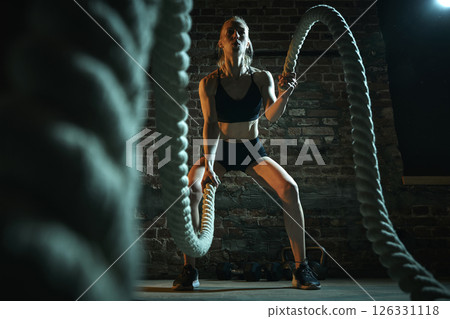 Woman slams heavy battle ropes in deep squat while generating symmetrical motion with explosive energy and sharp focus under gym spotlight. 126331118