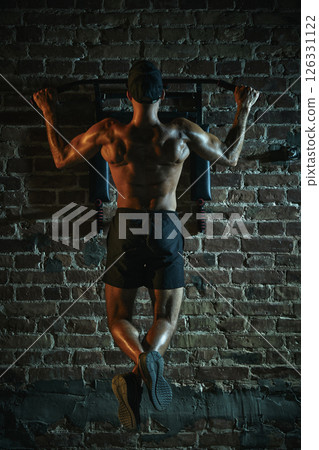 Rear view of man hanging on bar doing pull-up with muscular back, glistening under dramatic light, showcasing dedication and powerful physique. 126331122