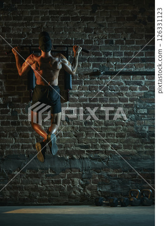 Rear view of muscular man performing pull-up from back view with defined upper body and crossed legs under spotlight in gritty gym atmosphere. 126331123
