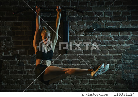 Woman lifting straight legs while hanging from bar in gym performing challenging core exercise with control, strength and balance. Woman lifting straight legs while hanging from bar in gym performing challenging core exercise with control, strength and balance. 126331124