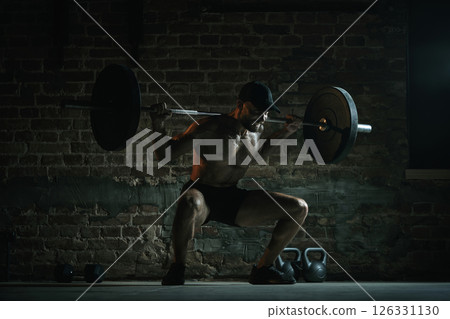 Shirtless muscular man performing deep barbell squat with loaded bar on shoulders under dramatic gym lighting with full intensity. 126331130