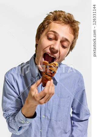 Man wearing striped shirt opening mouth to eat crispy croissant topped with chocolate glaze in joyful pose against white background. Man wearing striped shirt opening mouth to eat crispy croissant topped with chocolate glaze in joyful pose against white background. 126331144