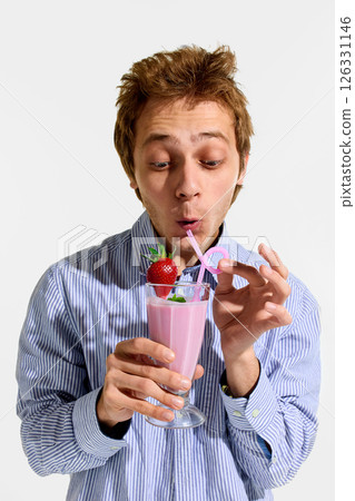 Young man sipping strawberry milkshake through pink curly straw, eyes wide open and expression excited while holding glass. Young man sipping strawberry milkshake through pink curly straw, eyes wide open and expression excited while holding glass. 126331146
