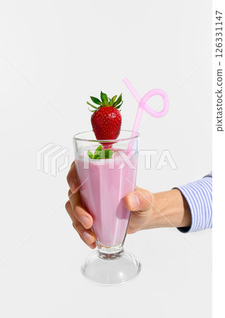 Hand holding tall glass of strawberry smoothie decorated with fresh strawberry, mint and pink curly straw against white studio background. Hand holding tall glass of strawberry smoothie decorated with fresh strawberry, mint and pink curly straw against white studio background. 126331147