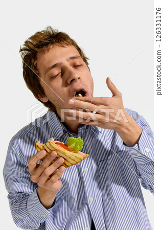 Playful man in striped shirt eating sandwich with eyes closed, savoring each bite with fingers near mouth and fresh lettuce inside sandwich. Playful man in striped shirt eating sandwich with eyes closed, savoring each bite with fingers near mouth and fresh lettuce inside sandwich. 126331176