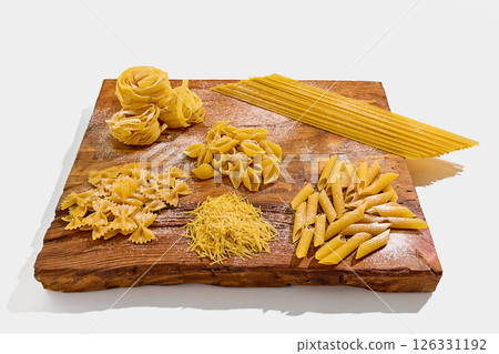 Various types of raw pasta neatly arranged on rustic wooden board covered in flour include penne, spaghetti, shells, farfalle, and fettuccine bundles. 126331192