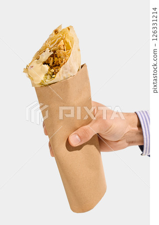 Shawarma roll with grilled meat, white sauce, cabbage, and flatbread held in brown paper sleeve, viewed from hand against white background. 126331214