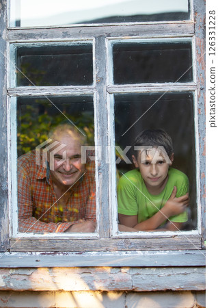 Grandfather and grandson looking out of the window of a village house and smiling. Father's Day. Boy and man in the window. Grandfather and grandson looking out of the window of a village house and smiling. Father's Day. Boy and man in the window. 126331228