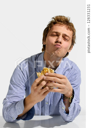 Young man in striped shirt closing eyes in satisfaction while chewing shawarma roll, hands holding wrap with visible sauce and filling. 126331231