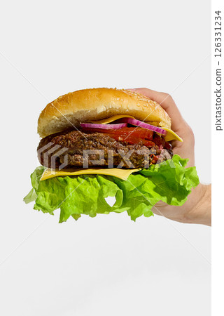 Close-up hand holding large burger with juicy grilled beef, cheddar cheese, onion rings, lettuce, and sesame bun against clean white background. 126331234