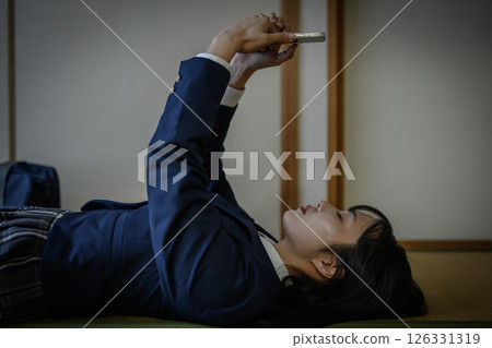 High school girl lying down in a Japanese-style room and looking at her smartphone: Bullying, school refusal, SNS troubles, social issues 126331319