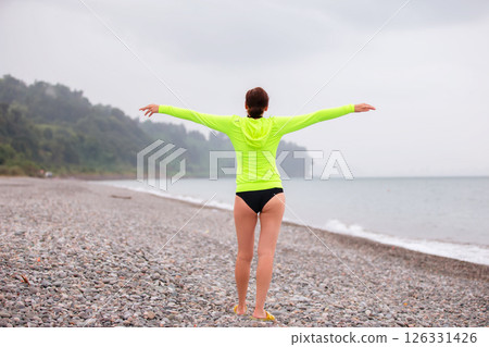 A full-length woman in a bright green sports jacket, her arms outstretched, looks out to sea. She stands with her back to the camera. 126331426