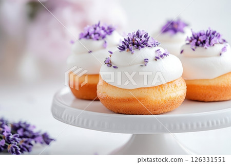 Delicate mini donuts topped with smooth white frosting and garnished with fresh lavender buds, presented on an elegant white ceramic cake stand.  Delicate mini donuts topped with smooth white frosting and garnished with fresh lavender buds, presented on an elegant white ceramic cake stand.  126331551