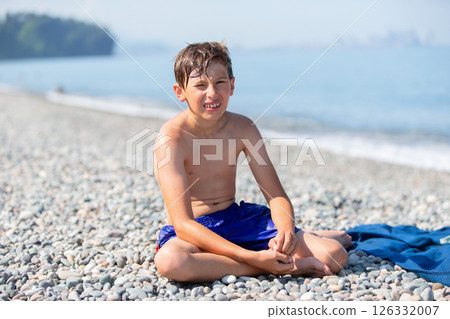 Funny teenage boy sitting on pebble beach and looking at camera. 126332007