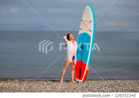 A woman stands on the beach with a SUP board. She is wearing a white shirt and blue swimming trunks. A woman stands on the beach with a SUP board. She is wearing a white shirt and blue swimming trunks. 126332248