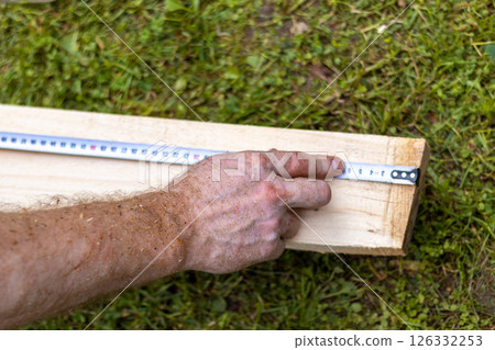 Man's hands measuring wooden board with tape measure on grass. Accuracy and precision of measurements. Outdoor setting emphasizes activity and DIY skills. Close up 126332253