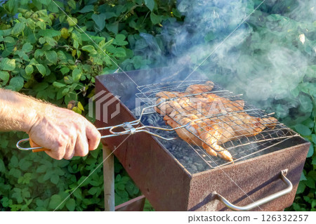 Grilling Chicken being grilled over charcoal in barbecue outdoors. Hand holds grill handle, highlighting cooking process amidst lush greenery and rising smoke Grilling Chicken being grilled over charcoal in barbecue outdoors. Hand holds grill handle, highlighting cooking process amidst lush greenery and rising smoke 126332257