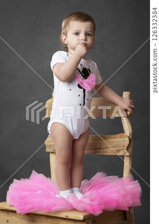 A little girl dresses in a pink tutu while standing on a wooden chair. She looks at the camera. 126332384