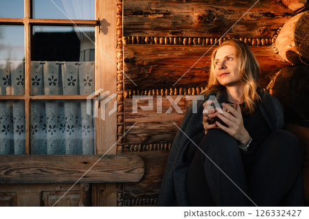 Woman wrapped in cozy blanket, savoring warm drink while sitting peacefully by window in rustic mountain chalet at sunset, enjoying tranquility of golden hour. Woman with tea in front of mountain hut 126332427