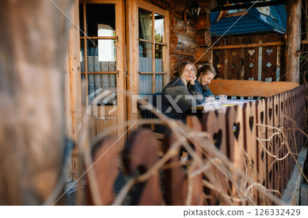 Two female friends are enjoying some relaxing time together, sitting at table on porch of traditional wooden chalet in mountain resort. Female friends relaxing together, sitting at wooden chalet porch 126332429