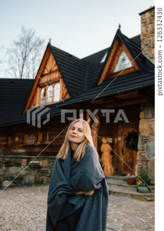 Blond woman wrapped in cozy gray blanket stands in front of charming wooden mountain chalet, enjoying peaceful holiday amidst serene countryside landscape. Blond woman standing before wooden chalet 126332430