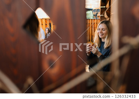 Young woman holding playing cards, smiling and peeking through wooden fence, enjoying card game with her friends in cozy outdoor setting. Female grasping playing cards, outdoor card game with friends 126332433