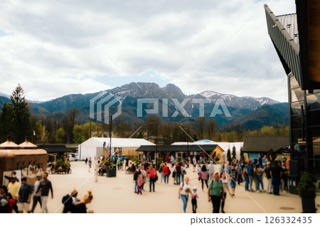 Tourists walking in mountain resort with wooden stalls, tents, and modern building, enjoying view of Tatra mountains with Giewont peak in Zakopane, Poland. Visitors stroll through mountain town 126332435