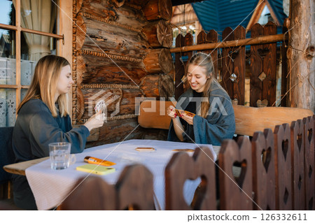 Two friends sitting at rustic wooden table on porch of cozy mountain chalet, playing cards and savoring their leisure time together in beautiful outdoors. Friends enjoying card game on mountain chalet 126332611