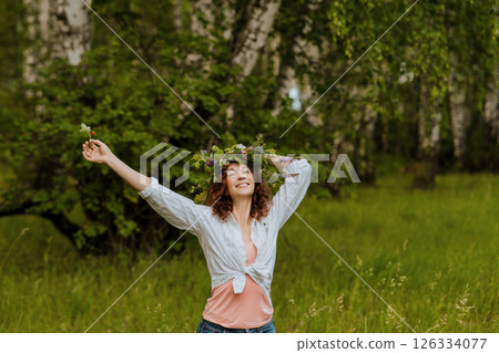 Joyful Caucasian woman wearing a flower wreath raises her arms in lush green forest during midsummer. The smiling adult celebrates Ivan Kupala surrounded by nature, symbolizing freedom and connection 126334077