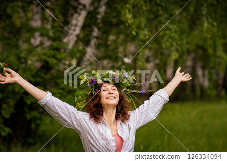 Smiling woman in white shirt and flower crown on green forest background. Joyful adult woman in a floral crown enjoying summer nature in a birch grove Smiling woman in white shirt and flower crown on green forest background. Joyful adult woman in a floral crown enjoying summer nature in a birch grove 126334094