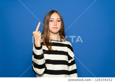 A Hispanic teenage girl about 15 years old wearing a striped sweater standing against a blue background, giving the middle finger. A rude and impolite expression, Fuck you. 126334252
