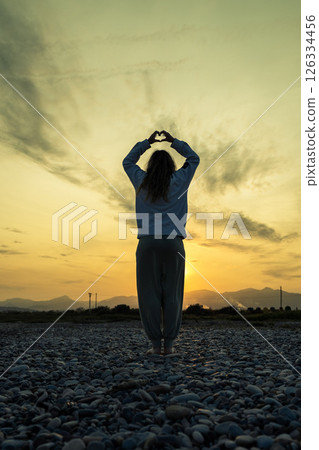 Woman creating heart shape at sunset on rocky beach 126334456