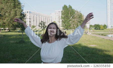 A joyful and carefree woman happily enjoying a sunny day in a vibrant green park with tall buildings in the background A joyful and carefree woman happily enjoying a sunny day in a vibrant green park with tall buildings in the background 126335850