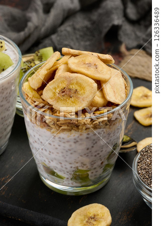 Two glasses of chia seed pudding, one topped with diced kiwi and the other with granola and banana chips, are on a dark surface with ingredients 126336489