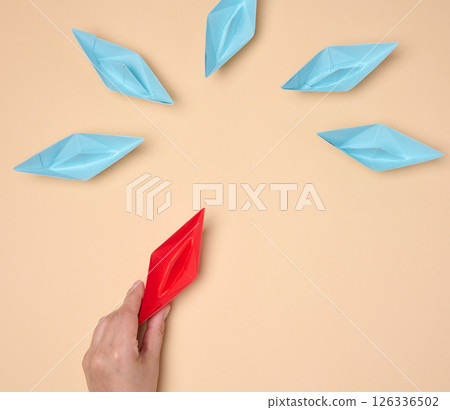 A hand holds a red origami paper boat next to a group of blue ones, symbolizing choice, uniqueness or a new idea 126336502