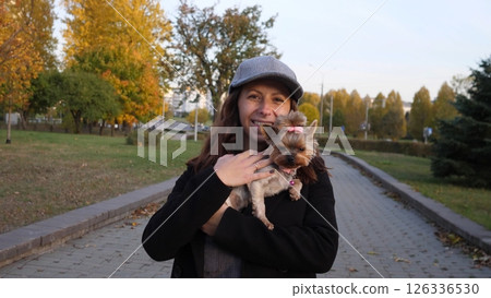 A joyful woman lovingly holds her small dog tightly among the vibrant, colorful autumn leaves in a peaceful park 126336530