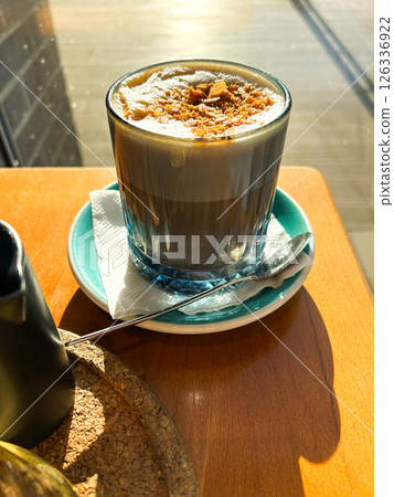 Cup of cappuccino with delicate foam art bathed in golden sunlight, placed on wooden table. Cozy atmosphere ideal for travel cafe lifestyle and social media content. Vertical. 126336922