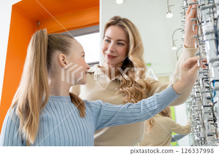 Mom and daughter choosing new eyeglasses in optic shop and looking involved Mom and daughter choosing new eyeglasses in optic shop and looking involved 126337998