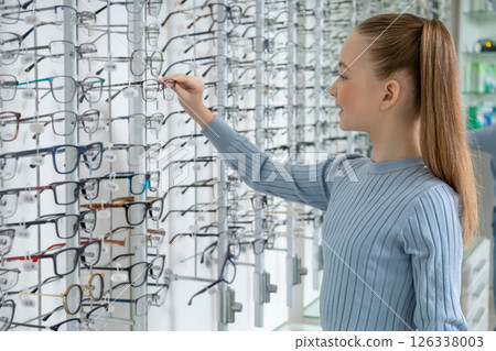 Long-haired caucasian girl choosing new eyeglasses in an optic shop 126338003