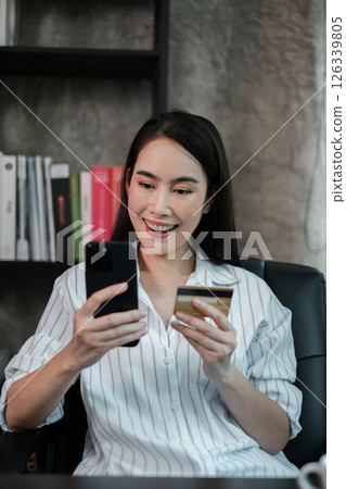 A cheerful woman using her smartphone and credit card for online shopping while sitting in a stylish home office. A cheerful woman using her smartphone and credit card for online shopping while sitting in a stylish home office. 126339805
