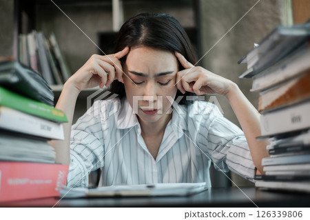 A businesswoman in a striped shirt appears stressed while sitting at a desk cluttered with stacks of documents and files. A businesswoman in a striped shirt appears stressed while sitting at a desk cluttered with stacks of documents and files. 126339806