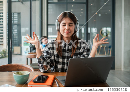 A young woman wearing headphones meditates at her desk with a laptop in a modern office setting, promoting relaxation and focus. 126339828